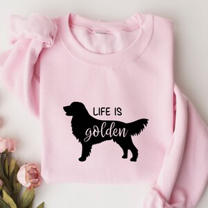 Pu&ograve; includere: Felpa rosa con una silhouette nera di un golden retriever e il testo "Life is golden".