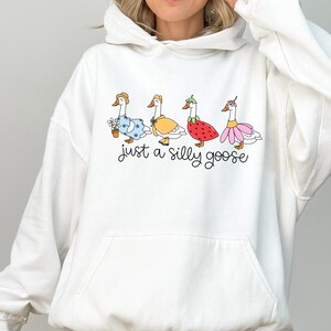 Peut inclure: Sweat-shirt &agrave; capuche blanc avec une illustration fantaisiste de quatre oies v&ecirc;tues de diff&eacute;rentes tenues. Les oies portent une robe bleue, un manteau jaune, un costume de fraise et un tutu rose. Le texte "just a silly goose" est &eacute;crit en dessous.
