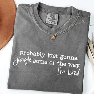 May include: A grey Comfort Colors t-shirt with the text "probably just gonna jingle some of the way I'm tired" in white script. A gold necklace with a pendant and a brown hat are also visible.