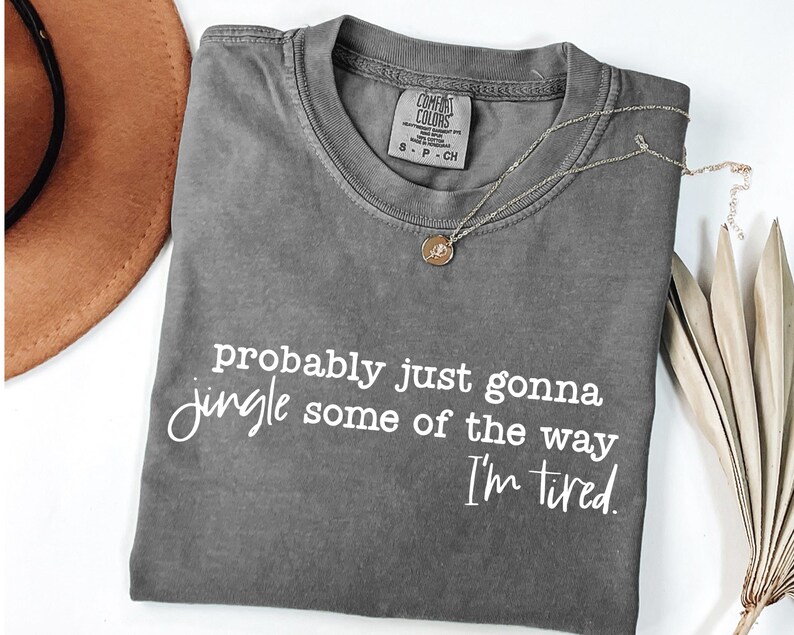 May include: A grey Comfort Colors t-shirt with the text "probably just gonna jingle some of the way I'm tired" in white script. A gold necklace with a pendant and a brown hat are also visible.