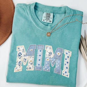 K&ouml;nnte beinhalten: Ein t&uuml;rkisfarbenes Comfort Colors T-Shirt mit dem Wort "MIMI" in einem Patchwork-Design aus Blumen-, Vichy- und Tupfenmustern appliziert. Eine goldene Halskette mit Anh&auml;nger liegt auf dem Shirt. Das Shirt ist gefaltet.
