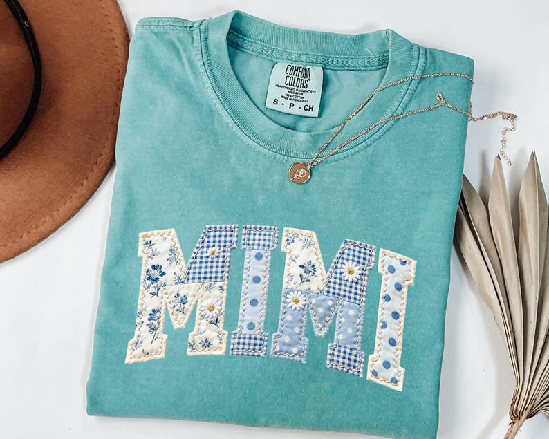 K&ouml;nnte beinhalten: Ein t&uuml;rkisfarbenes Comfort Colors T-Shirt mit dem Wort "MIMI" in einem Patchwork-Design aus Blumen-, Vichy- und Tupfenmustern appliziert. Eine goldene Halskette mit Anh&auml;nger liegt auf dem Shirt. Das Shirt ist gefaltet.