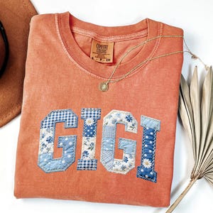 May include: An orange Comfort Colors t-shirt with the name "GIGI" appliqued in blue and white patchwork fabric. A gold necklace with a pendant rests on the shirt. A brown hat and dried palm leaf are in the background.