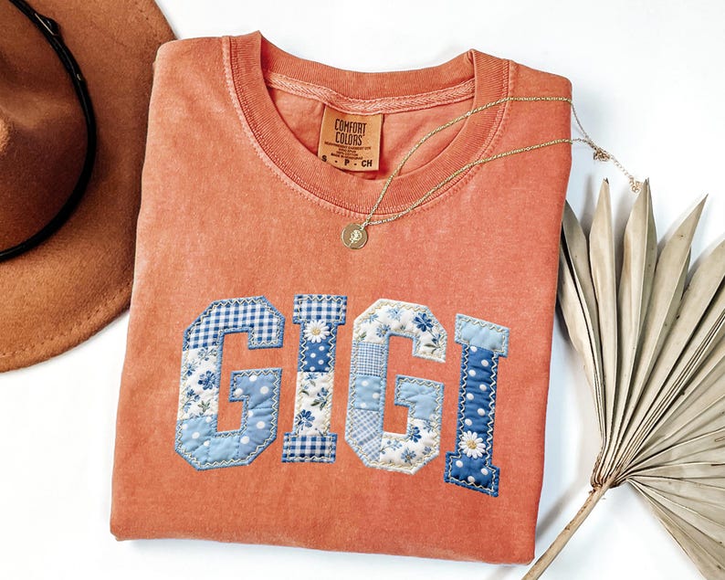May include: An orange Comfort Colors t-shirt with the name "GIGI" appliqued in blue and white patchwork fabric. A gold necklace with a pendant rests on the shirt. A brown hat and dried palm leaf are in the background.