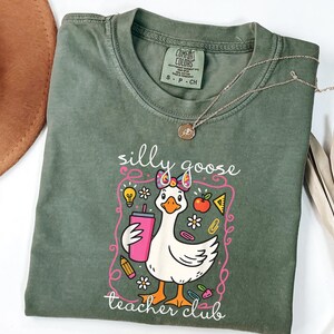 May include: A sage green t-shirt with a graphic of a cartoon goose holding a pink tumbler. The goose has a bow and is surrounded by school-themed illustrations. The text "silly goose teacher club" is printed below the goose.