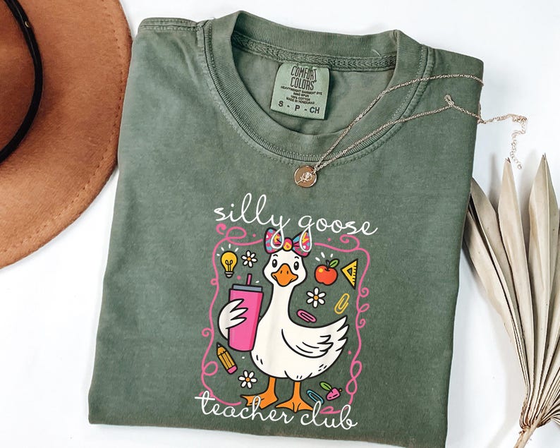 May include: A sage green t-shirt with a graphic of a cartoon goose holding a pink tumbler. The goose has a bow and is surrounded by school-themed illustrations. The text "silly goose teacher club" is printed below the goose.