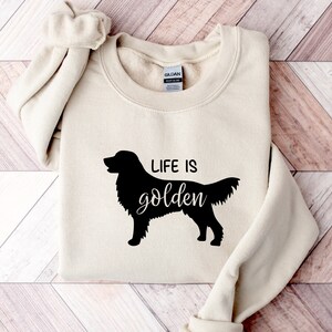 Pu&ograve; includere: Una felpa beige con una silhouette nera di un golden retriever e il testo "Life is golden".