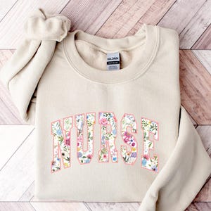 May include: A beige sweatshirt with the word "NURSE" printed in large, pink letters. The letters are filled with a floral pattern.