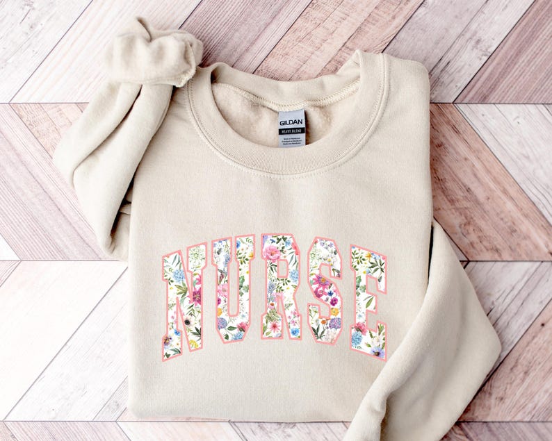 May include: A beige sweatshirt with the word "NURSE" printed in large, pink letters. The letters are filled with a floral pattern.