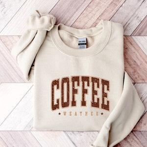 Puede incluir: Una sudadera beige con el texto "COFFEE WEATHER" en letras marrones con un aspecto desgastado. El texto est&aacute; en una fuente de estilo vintage.