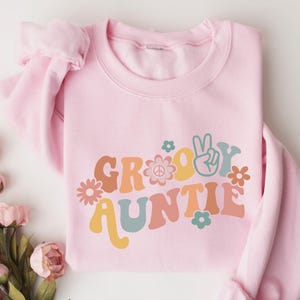 May include: Pink sweatshirt with a groovy auntie graphic in retro colors and a peace sign. The graphic includes flowers and a peace sign.