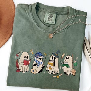 May include: Sage green Comfort Colors t-shirt featuring a whimsical design of four cartoon ghosts, each with a book or magical item. The ghosts are in a row, with a gold necklace draped across the shirt.