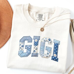 May include: Cream-colored t-shirt with the word "GIGI" appliqu&eacute;d in blue patchwork fabric. The letters feature various patterns, including gingham and floral prints. A gold necklace with a pendant and a brown hat are also visible.