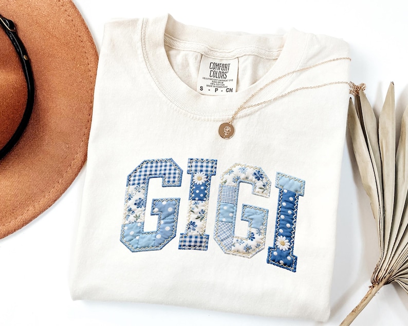 May include: Cream-colored t-shirt with the word "GIGI" appliqu&eacute;d in blue patchwork fabric. The letters feature various patterns, including gingham and floral prints. A gold necklace with a pendant and a brown hat are also visible.