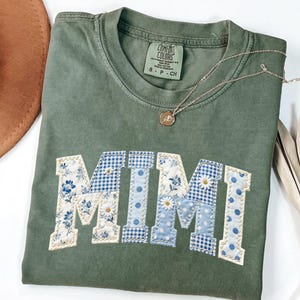 K&ouml;nnte beinhalten: Olivgr&uuml;nes Comfort Colors T-Shirt mit dem Wort "MIMI" in einem Patchwork-Design appliziert. Die Buchstaben zeigen Blumen-, Vichy- und Tupfenmuster in Blau und Wei&szlig;. Eine goldene Halskette mit Anh&auml;nger liegt auf dem Shirt.