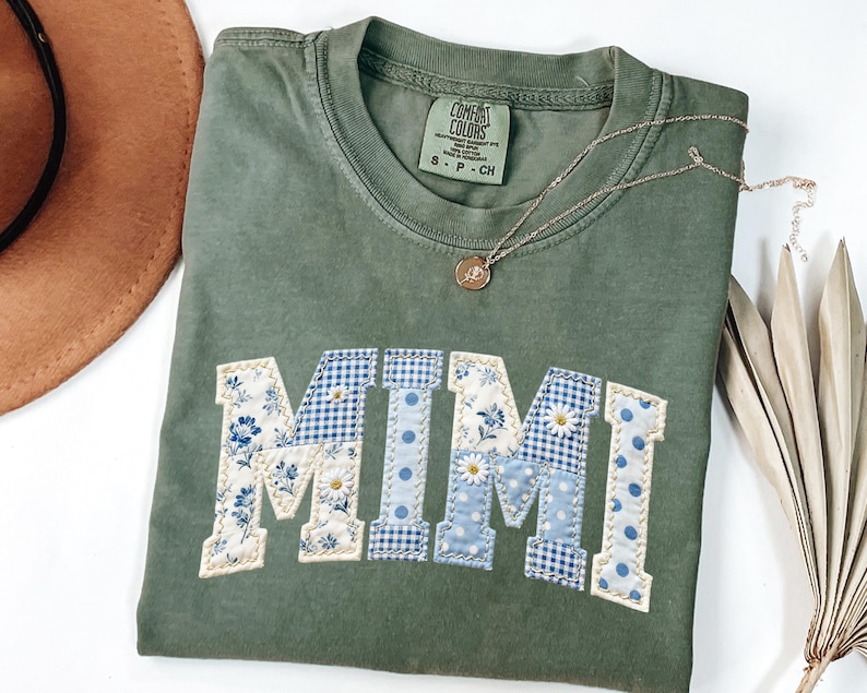 K&ouml;nnte beinhalten: Olivgr&uuml;nes Comfort Colors T-Shirt mit dem Wort "MIMI" in einem Patchwork-Design appliziert. Die Buchstaben zeigen Blumen-, Vichy- und Tupfenmuster in Blau und Wei&szlig;. Eine goldene Halskette mit Anh&auml;nger liegt auf dem Shirt.