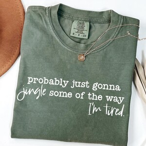 May include: An olive green t-shirt with the text "probably just gonna jingle some of the way I'm tired" in white script. A gold necklace with a pendant rests on the shirt. A brown hat is visible in the upper left corner.