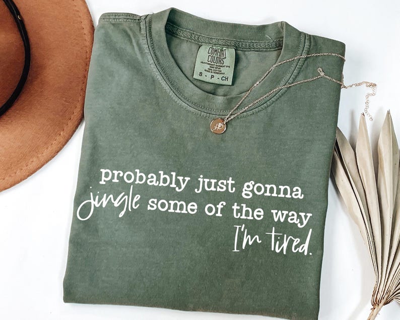 May include: An olive green t-shirt with the text "probably just gonna jingle some of the way I'm tired" in white script. A gold necklace with a pendant rests on the shirt. A brown hat is visible in the upper left corner.