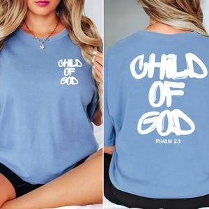 May include: A light blue t-shirt with the words "CHILD OF GOD" printed in white on the front and back. The back of the shirt also includes "PSALM 23" in white. The shirt is a comfortable, casual style.