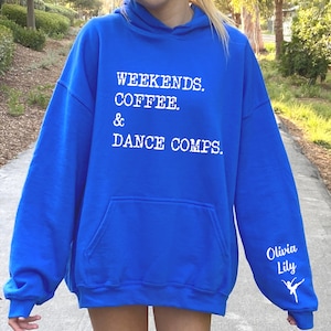 Op de afbeelding: Koninklijk blauwe hoodie met witte tekst die "Weekends. Coffee. & Dance Comps." luidt en een wit danser silhouet op de mouw met de tekst "Olivia Lily".