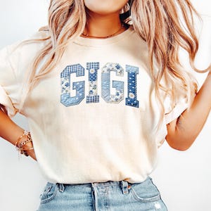May include: Cream-colored t-shirt with the word "GIGI" in blue patchwork letters. The letters feature floral, gingham, and solid blue patterns. The shirt is a casual, everyday wear item.