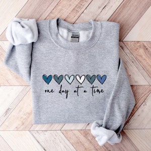 May include: Gray sweatshirt with a row of blue, teal, and white hearts and the text "one day at a time" printed on the front.