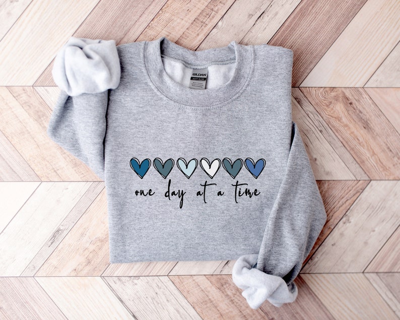 May include: Gray sweatshirt with a row of blue, teal, and white hearts and the text "one day at a time" printed on the front.