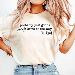 May include: A cream-coloured t-shirt featuring the text "probably just gonna jingle some of the way I'm tired" in black script. The shirt is paired with blue jeans. The image depicts casual clothing.