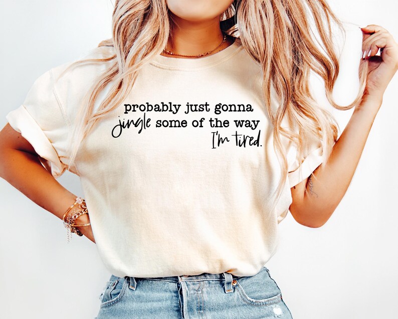 May include: A cream-coloured t-shirt featuring the text "probably just gonna jingle some of the way I'm tired" in black script. The shirt is paired with blue jeans. The image depicts casual clothing.