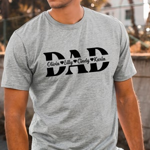 May include: Gray t-shirt with the word "DAD" in large black letters.  The names Olivia, Lilly, Cindy, and Karla are written in smaller black letters below the word "DAD" with a heart symbol between each name.