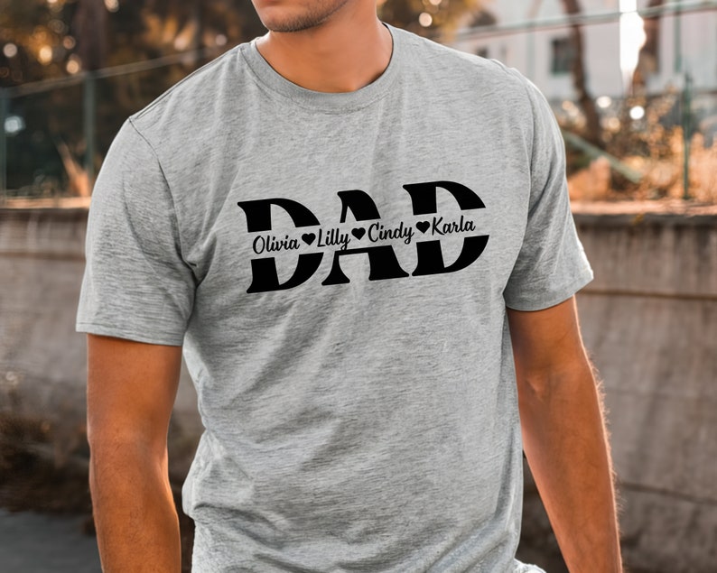May include: Gray t-shirt with the word "DAD" in large black letters.  The names Olivia, Lilly, Cindy, and Karla are written in smaller black letters below the word "DAD" with a heart symbol between each name.