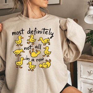 May include: A beige sweatshirt with the phrase "most definitely not all in a row" in black text. The sweatshirt features cartoon yellow duck illustrations. The ducks are in various poses, some walking and some lying down.