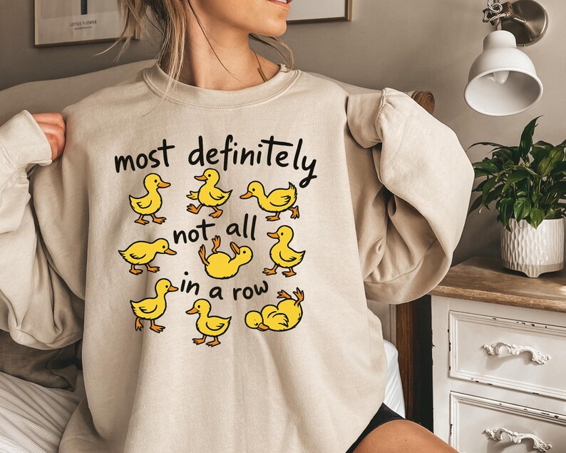May include: A beige sweatshirt with the phrase "most definitely not all in a row" in black text. The sweatshirt features cartoon yellow duck illustrations. The ducks are in various poses, some walking and some lying down.