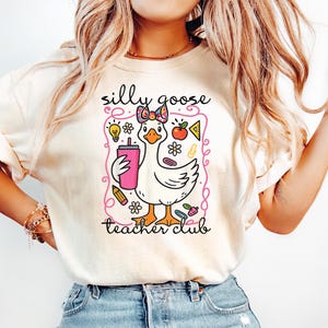May include: Cream-colored t-shirt featuring a cartoon goose holding a pink drink. The goose has a bow and is surrounded by school-themed graphics. Text on the shirt reads "silly goose teacher club."