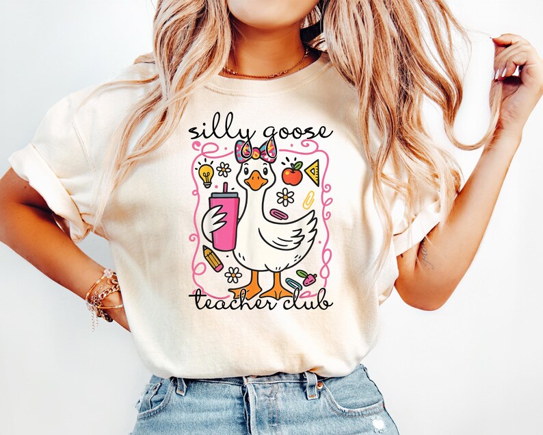 May include: Cream-colored t-shirt featuring a cartoon goose holding a pink drink. The goose has a bow and is surrounded by school-themed graphics. Text on the shirt reads "silly goose teacher club."