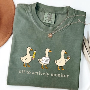 May include: An olive green crew neck t-shirt featuring a graphic of three light-colored ducks. The first duck holds a yellow pencil, the second wears a white t-shirt with the text "YOU GOT THIS," and the third holds a coffee cup. The text "off to actively monitor" is printed below the ducks.