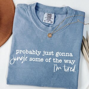 May include: A blue Comfort Colors t-shirt with the text "probably just gonna jingle some of the way I'm tired" in white. A gold necklace with a pendant and a brown hat are also visible.