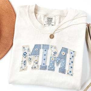 K&ouml;nnte beinhalten: Cremefarbenes T-Shirt mit dem Wort "MIMI" in blauem Blumen-, Vichy- und Tupfenmuster appliziert. Eine goldene Halskette mit Anh&auml;nger liegt auf dem Shirt. Ein brauner Hut und eine getrocknete Pflanze befinden sich im Hintergrund.