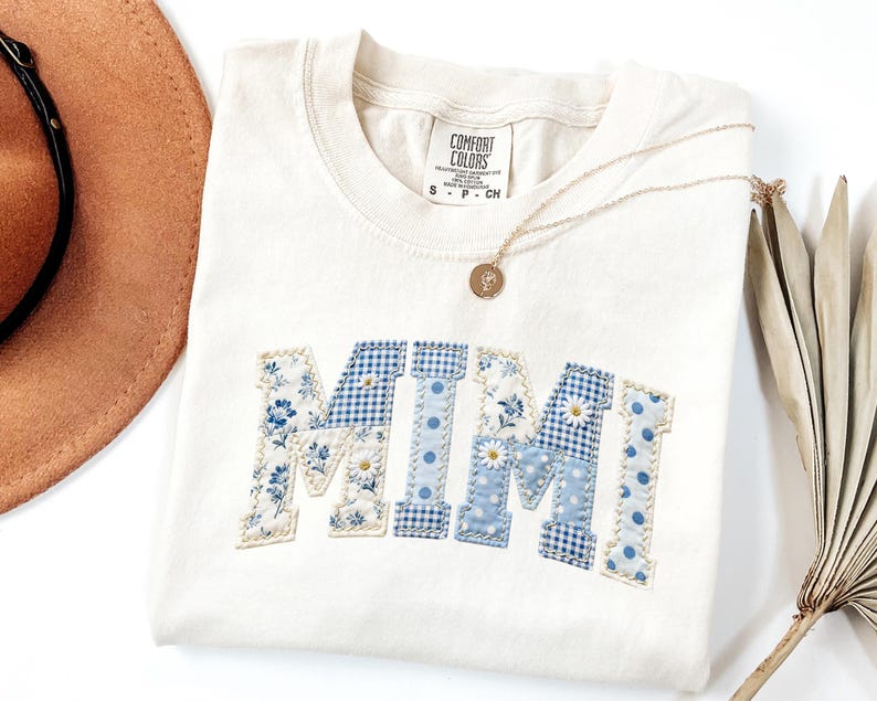 K&ouml;nnte beinhalten: Cremefarbenes T-Shirt mit dem Wort "MIMI" in blauem Blumen-, Vichy- und Tupfenmuster appliziert. Eine goldene Halskette mit Anh&auml;nger liegt auf dem Shirt. Ein brauner Hut und eine getrocknete Pflanze befinden sich im Hintergrund.
