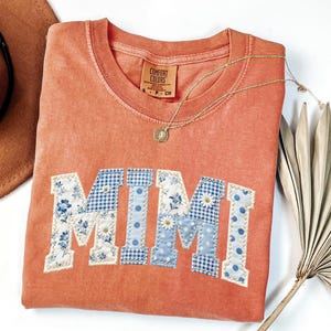 K&ouml;nnte beinhalten: Korallenfarbenes Comfort Colors Sweatshirt mit dem Wort "MIMI" in einem Patchwork-Design aus Blumen-, Vichy- und Tupfenstoffen. Eine goldene Halskette mit Anh&auml;nger liegt auf dem Shirt. Ein brauner Hut und ein getrockneter Palmwedel sind im Hintergrund.