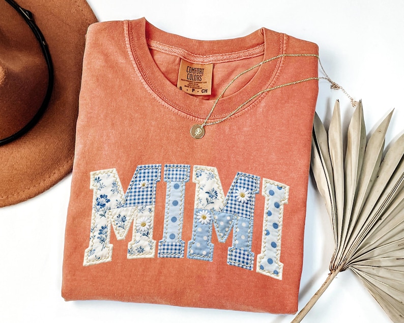 K&ouml;nnte beinhalten: Korallenfarbenes Comfort Colors Sweatshirt mit dem Wort "MIMI" in einem Patchwork-Design aus Blumen-, Vichy- und Tupfenstoffen. Eine goldene Halskette mit Anh&auml;nger liegt auf dem Shirt. Ein brauner Hut und ein getrockneter Palmwedel sind im Hintergrund.