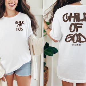 May include: White t-shirt with the text "CHILD OF GOD" in brown on the front and back. The back of the shirt also includes "PSALM 23" in brown. The shirt is a casual, oversized fit.