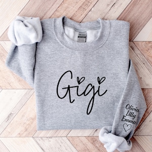 May include: Gray sweatshirt with the word "Gigi" in black cursive lettering and two small hearts above the word. The right sleeve has the names "Olivia, Lilly, Emma" in black lettering with a small heart below the names.