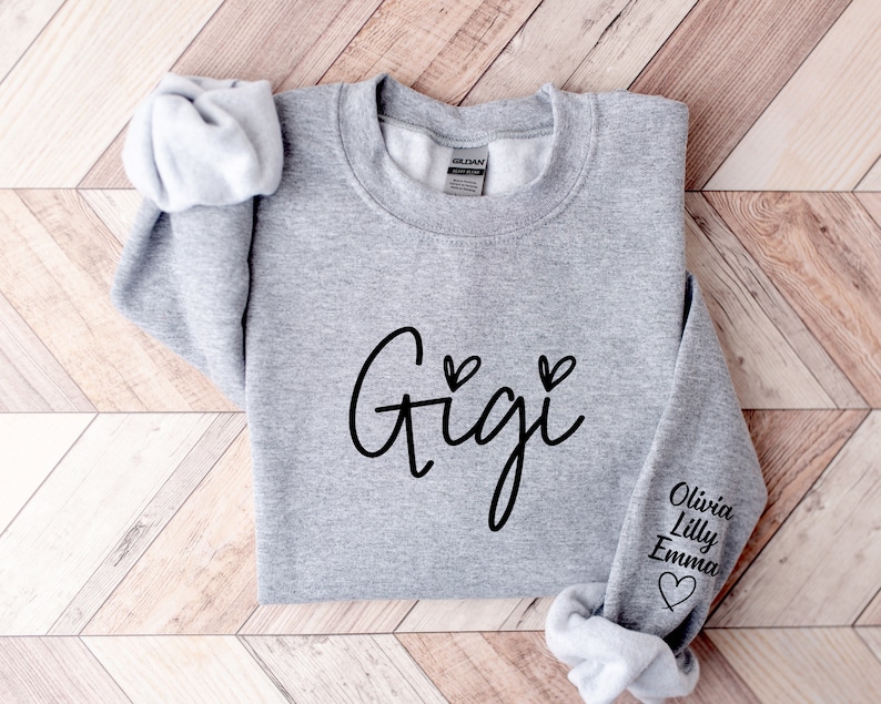 May include: Gray sweatshirt with the word "Gigi" in black cursive lettering and two small hearts above the word. The right sleeve has the names "Olivia, Lilly, Emma" in black lettering with a small heart below the names.