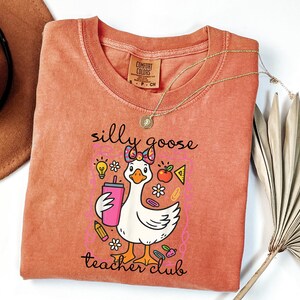 May include: A coral-colored Comfort Colors t-shirt featuring a cartoon goose with a bow, holding a pink drink. The shirt has the text "silly goose teacher club" and includes school-themed graphics. A gold necklace is draped across the shirt.