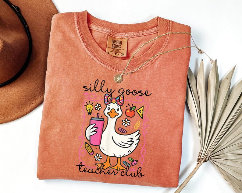 May include: A coral-colored Comfort Colors t-shirt featuring a cartoon goose with a bow, holding a pink drink. The shirt has the text "silly goose teacher club" and includes school-themed graphics. A gold necklace is draped across the shirt.