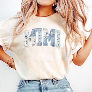 K&ouml;nnte beinhalten: Cremefarbenes T-Shirt mit dem Wort "MIMI" in blauen Patchwork-Buchstaben. Die Buchstaben weisen Blumen-, Vichy- und Tupfenmuster auf. Das Shirt hat kurze &Auml;rmel und eine lockere Passform.
