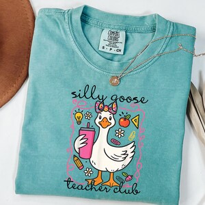 May include: A teal Comfort Colors t-shirt featuring a cartoon goose with a pink bow, holding a pink tumbler. The shirt has the text "silly goose teacher club" in a whimsical font, with school-themed graphics.