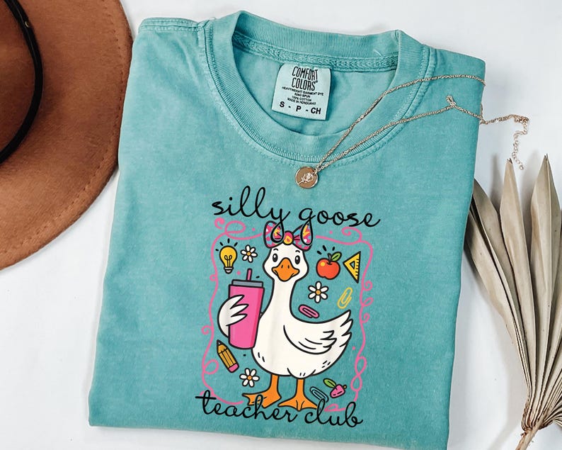 May include: A teal Comfort Colors t-shirt featuring a cartoon goose with a pink bow, holding a pink tumbler. The shirt has the text "silly goose teacher club" in a whimsical font, with school-themed graphics.
