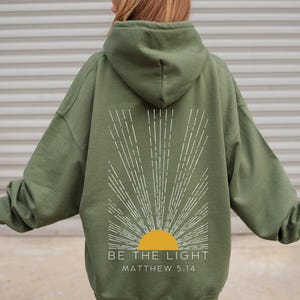 May include: A green hooded sweatshirt with a white sunburst design and the text "BE THE LIGHT" and "MATTHEW 5:14" printed on the back.